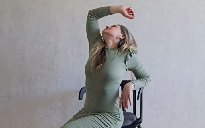 A woman in a long-sleeved dress poses artistically on a chair indoors, showcasing elegance.